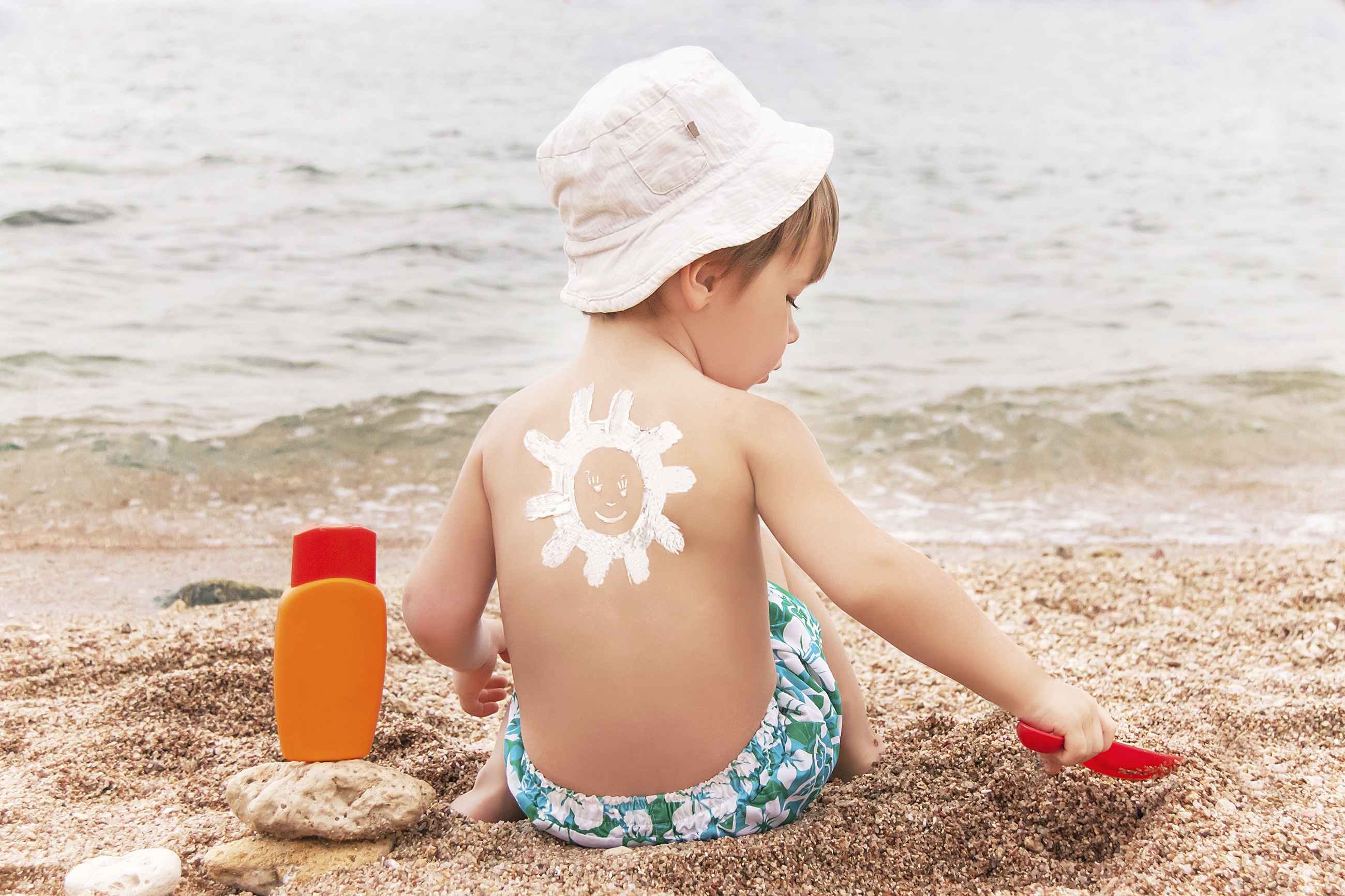 The sun drawing sunscreen (suntan lotion) on baby (boy)  back. Caucasian child is sitting with plastic container of sunscreen and toy on sunny beach. Close up, outdoor (Sharm El Sheikh, Egypt).